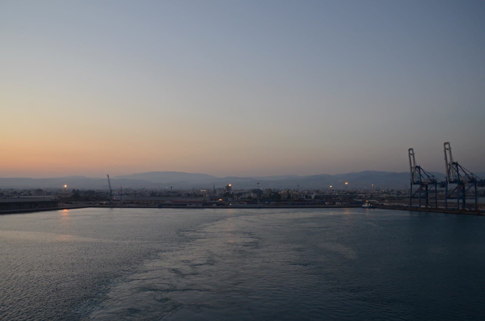 The port of Limassol at dusk, with harbour cranes and the city skyline silhouetted against a fading orange and blue sky over calm water